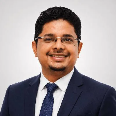 Professional headshot of a man wearing a navy suit, white shirt, and blue tie, smiling at the camera with a light gray studio background.